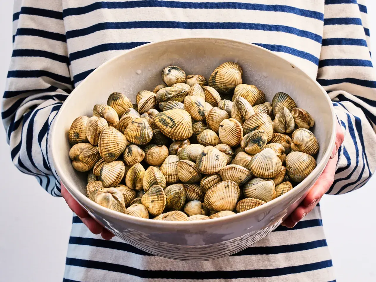Un bol rempli de coques coquillage, prêt à être cuisiné. Les coquillages sont disposés dans un bol en céramique, tenus par une personne portant un pull rayé bleu et blanc.