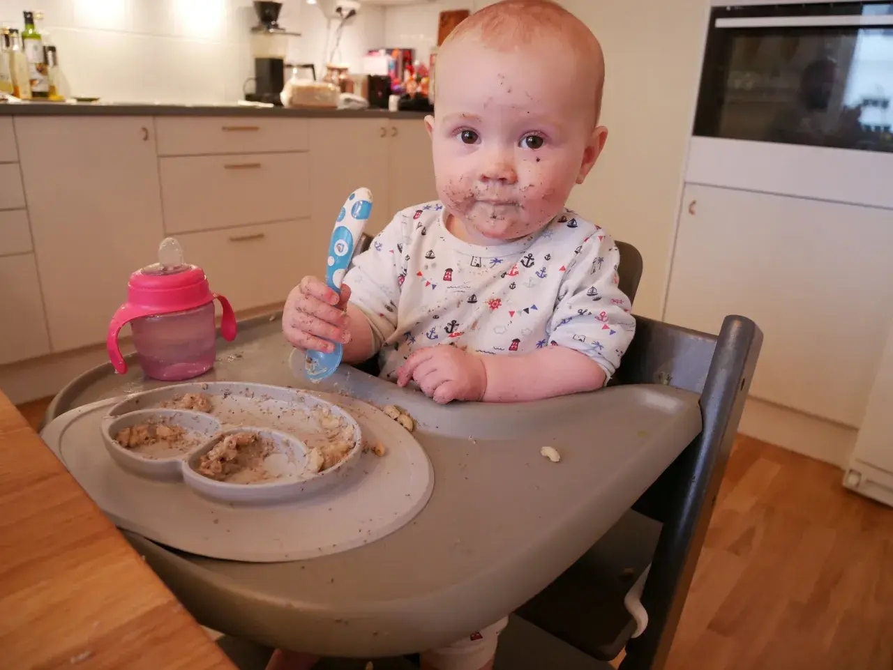 Sött litet barn med mat i ansiktet, redo att introducera mat bebis. En tallrik med mat och en mugg finns bredvid.