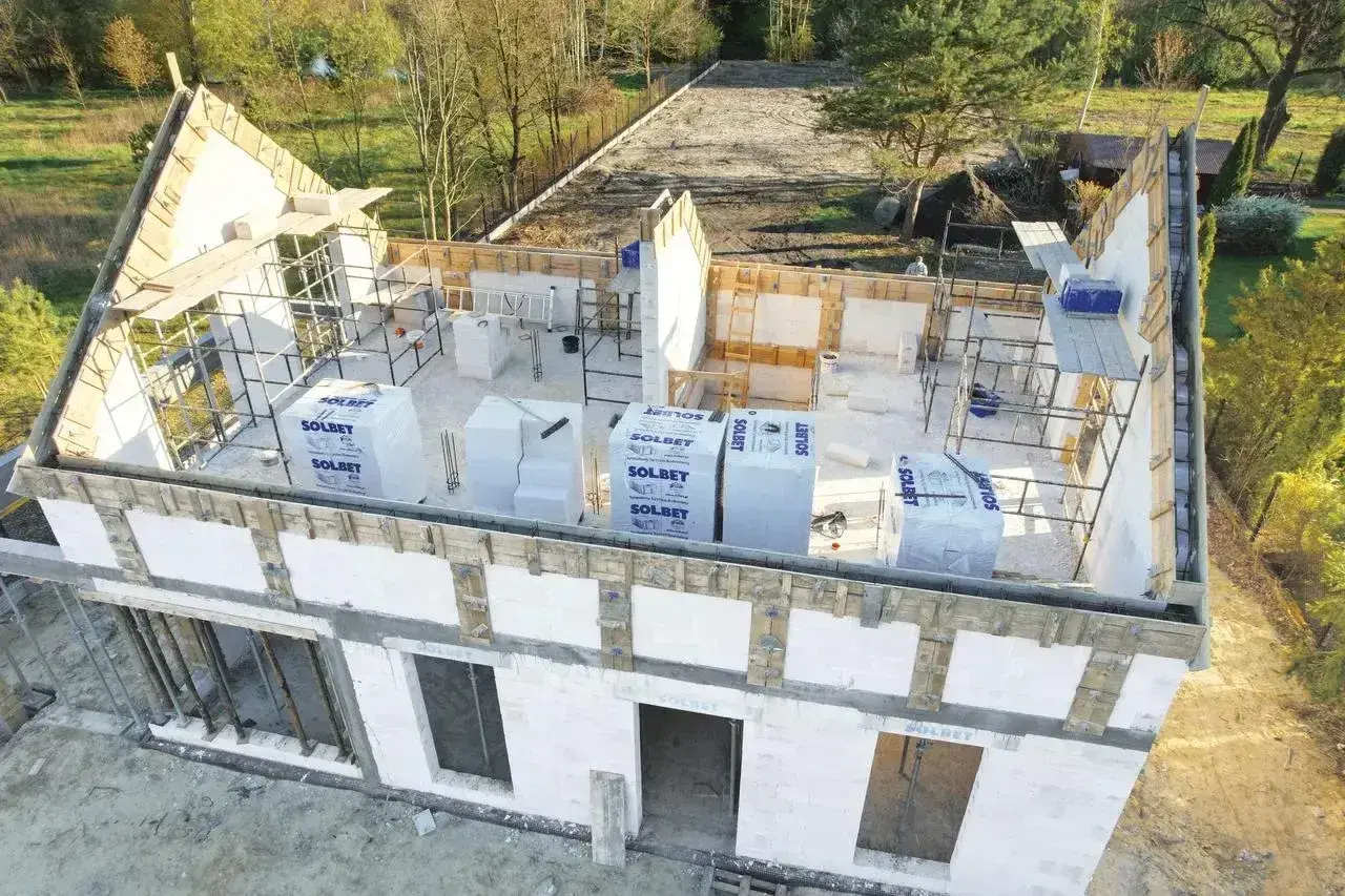 Budowa domu w toku, widoczne szalunki i materiały budowlane. Widać charakterystyczne trójkątne **goble** dachu, które czekają na pokrycie.