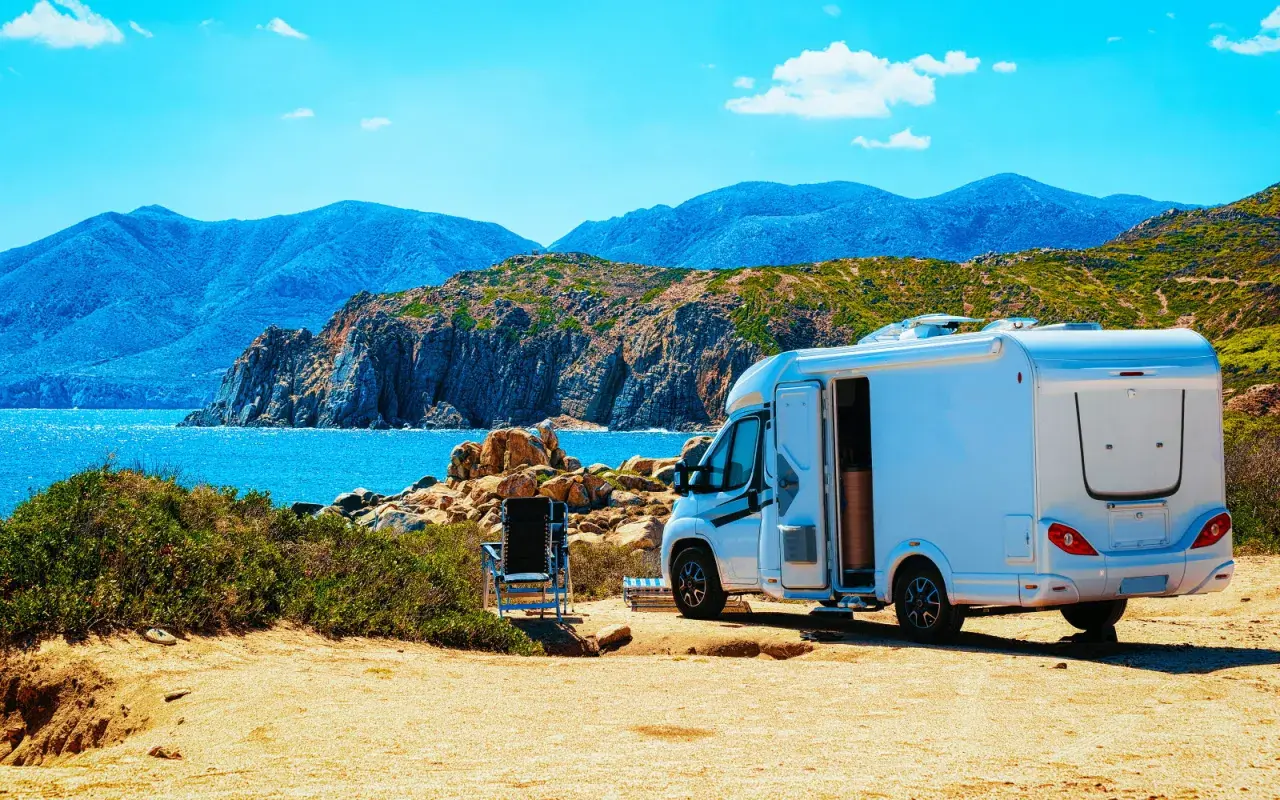 Un camping-car blanc est garé sur un terrain sablonneux, face à une mer bleue et des montagnes. L'application spot camping car idéale pour ce paysage.
