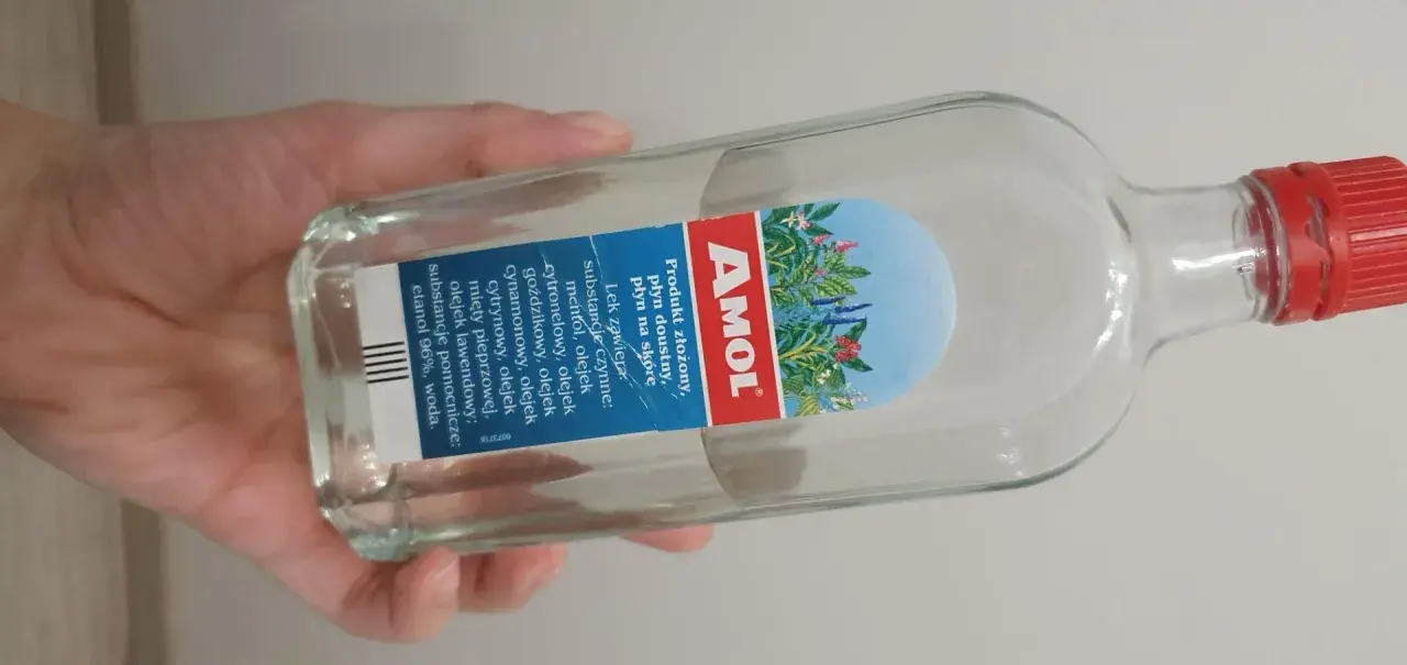 Dłoń trzyma butelkę Amolu. Ten lek, znany z dobrej ceny, zawiera mieszankę olejków eterycznych.
