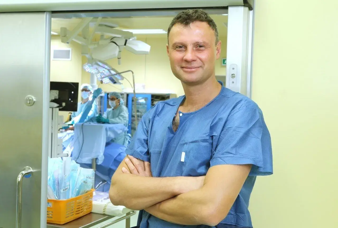 Lekarz w niebieskiej koszuli chirurgicznej stoi na tle sali operacyjnej.