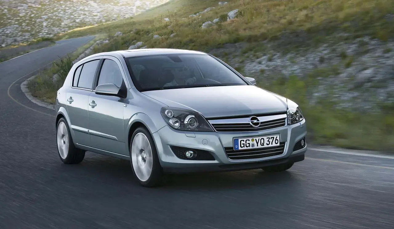 Srebrny Opel Astra 2005 na krętej drodze, otoczony zielonymi wzgórzami.
