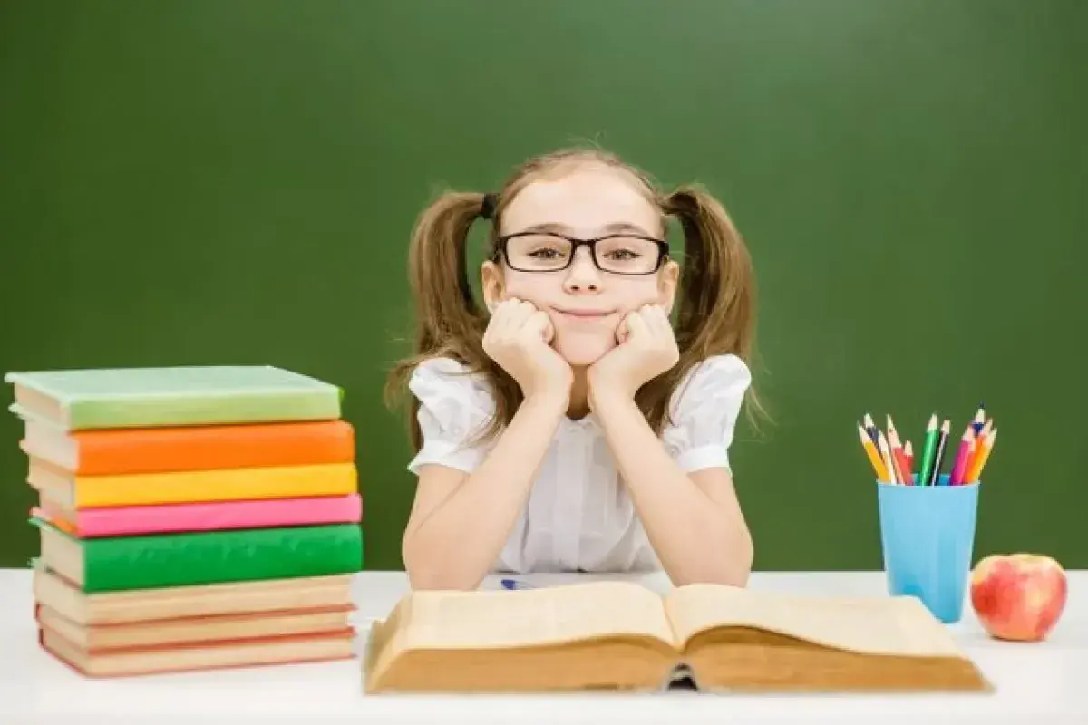 Uśmiechnięta dziewczynka w okularach z kitkami, oparta o ręce, siedzi przy biurku z książkami i jabłkiem. To idealny ipet do nauki.
