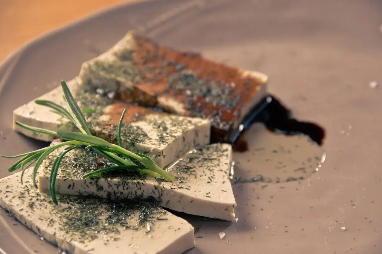 Queso tofu marinado con hierbas y romero, servido en un plato con salsa oscura.