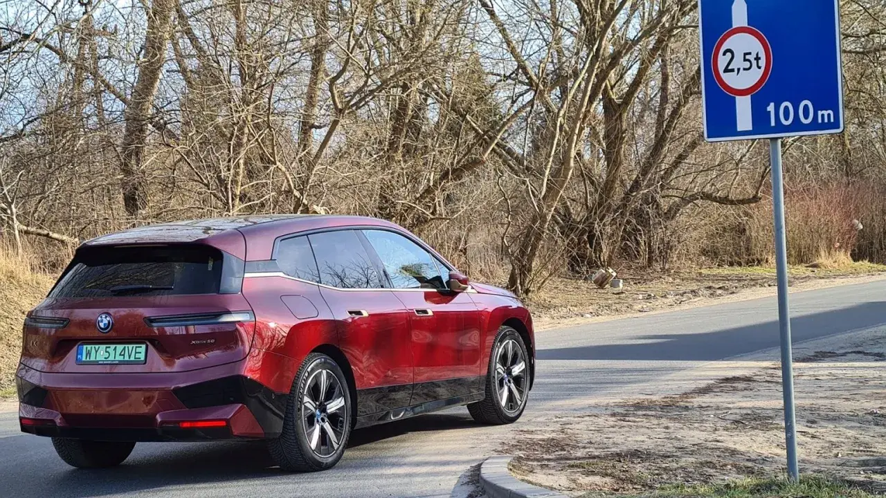 Czerwony BMW iX jedzie drogą, zbliżając się do znaku informującego o zakazie tonażowym 2,5 tony na dystansie 100m.