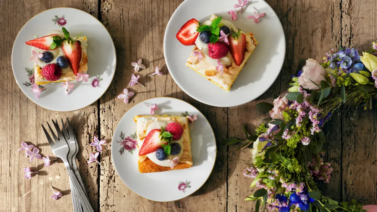 Tre tallrikar med vegansk midsommarmat, toppad med bär och blommor, ligger på ett träbord. En blomsterkrans ligger bredvid.