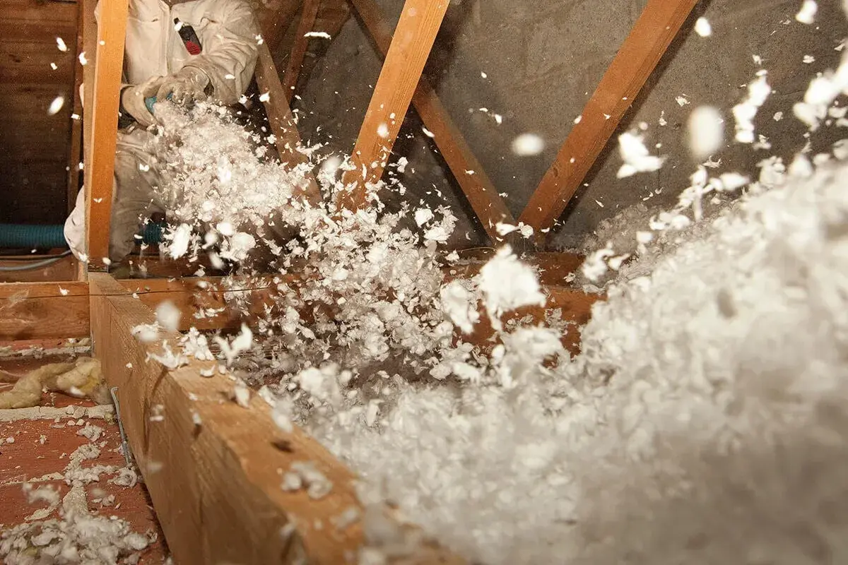 Un ouvrier en combinaison souffle de la laine de verre dans un grenier, créant un nuage blanc et cotonneux pour une isolation efficace.