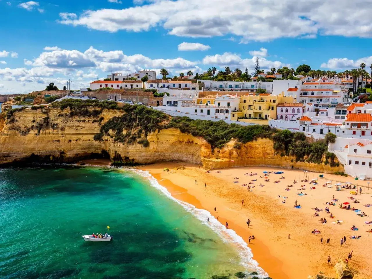 Sonnige Küste Portugals im Oktober. Ein Boot auf dem türkisfarbenen Meer vor einer Bucht mit Sandstrand und weiß-gelben Häusern.