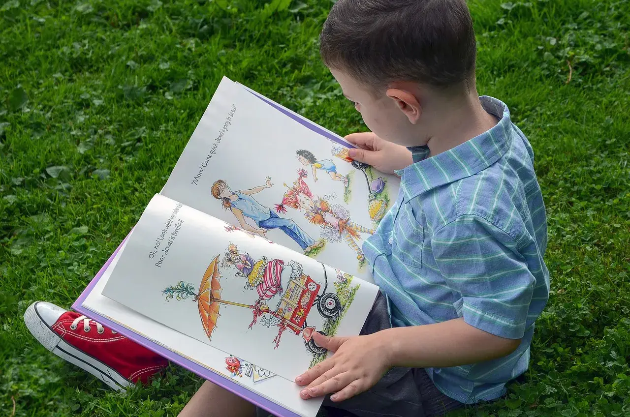 Chłopiec w niebieskiej koszuli czyta otwartą książkę z kolorowymi ilustracjami na trawie.