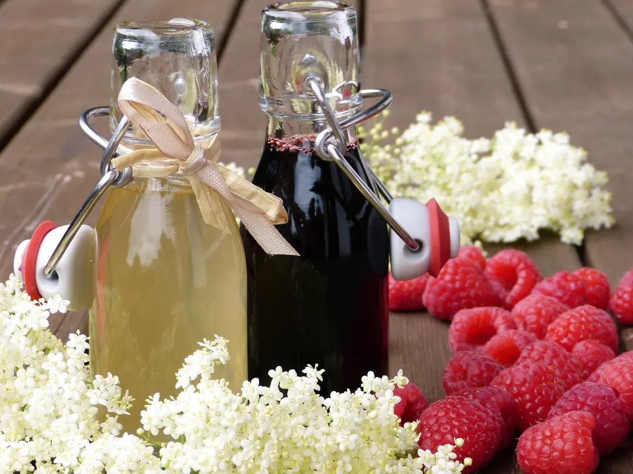 Zwei Flaschen mit selbstgemachtem Sirup, umgeben von Holunderblüten und Himbeeren. Perfekt zum Sirup einkochen.