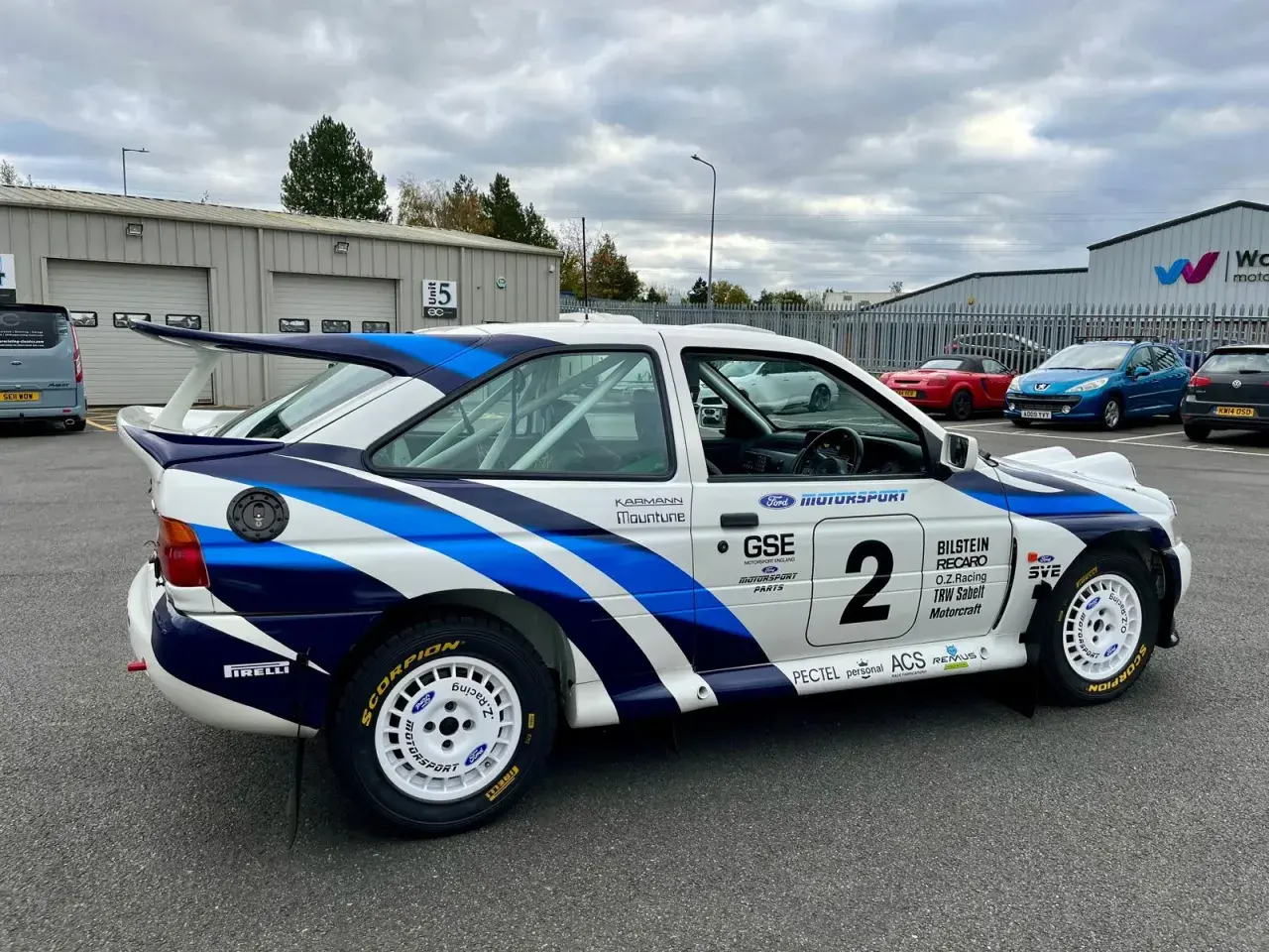 En vit rallybil Ford Escort Cosworth med blåa detaljer och nummer 2. Bilen står parkerad utomhus.