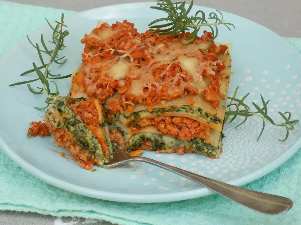 Pyszny przepis na lasagne z mięsem mielonym i szpinakiem, podana na talerzu z gałązkami rozmarynu.