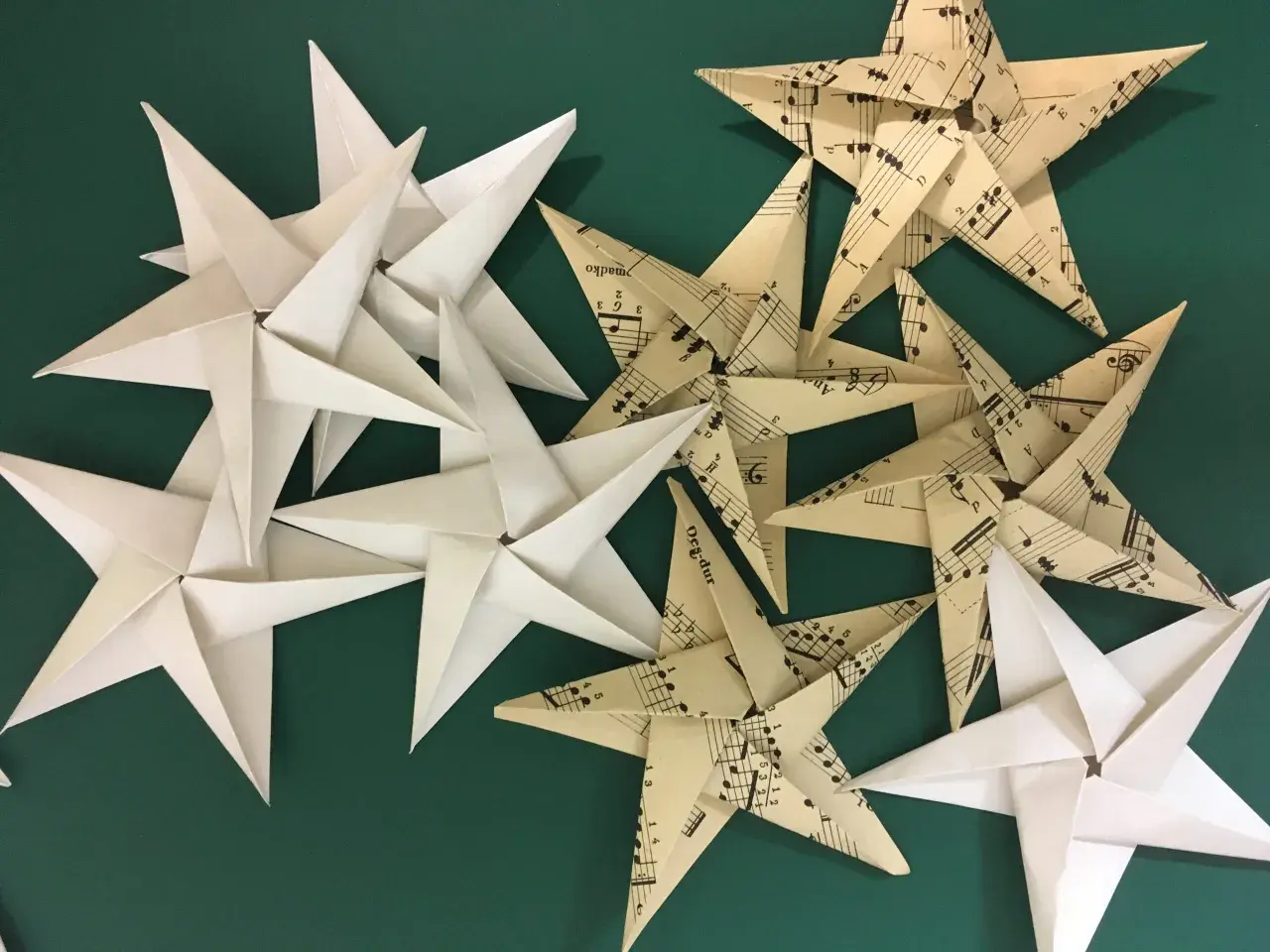 Gwiazdki z papieru, jedne białe, drugie z nutami, tworzą świąteczną kompozycję na zielonym tle.