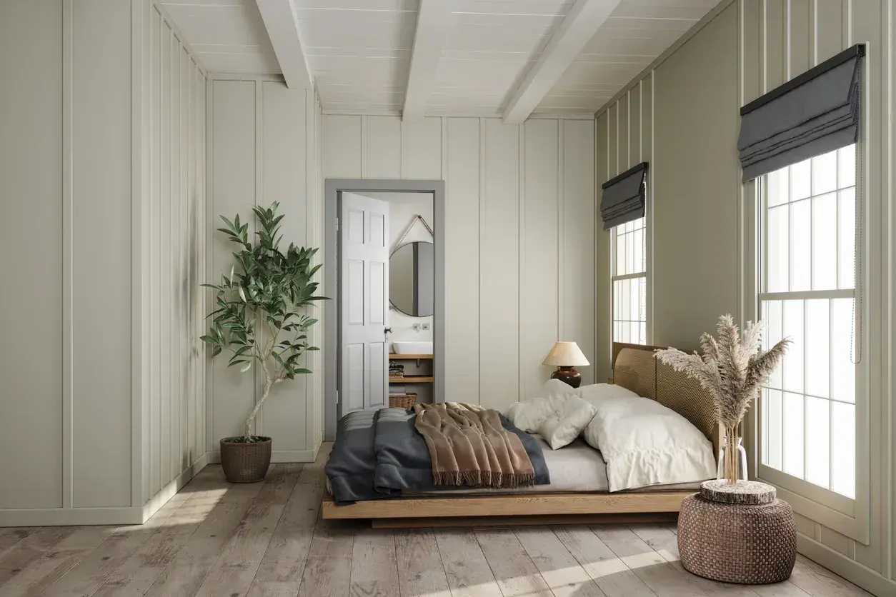 Letto in legno con coperte, posizionato di fianco alla porta aperta che conduce al bagno. Pianta in vaso e finestra con luce naturale.