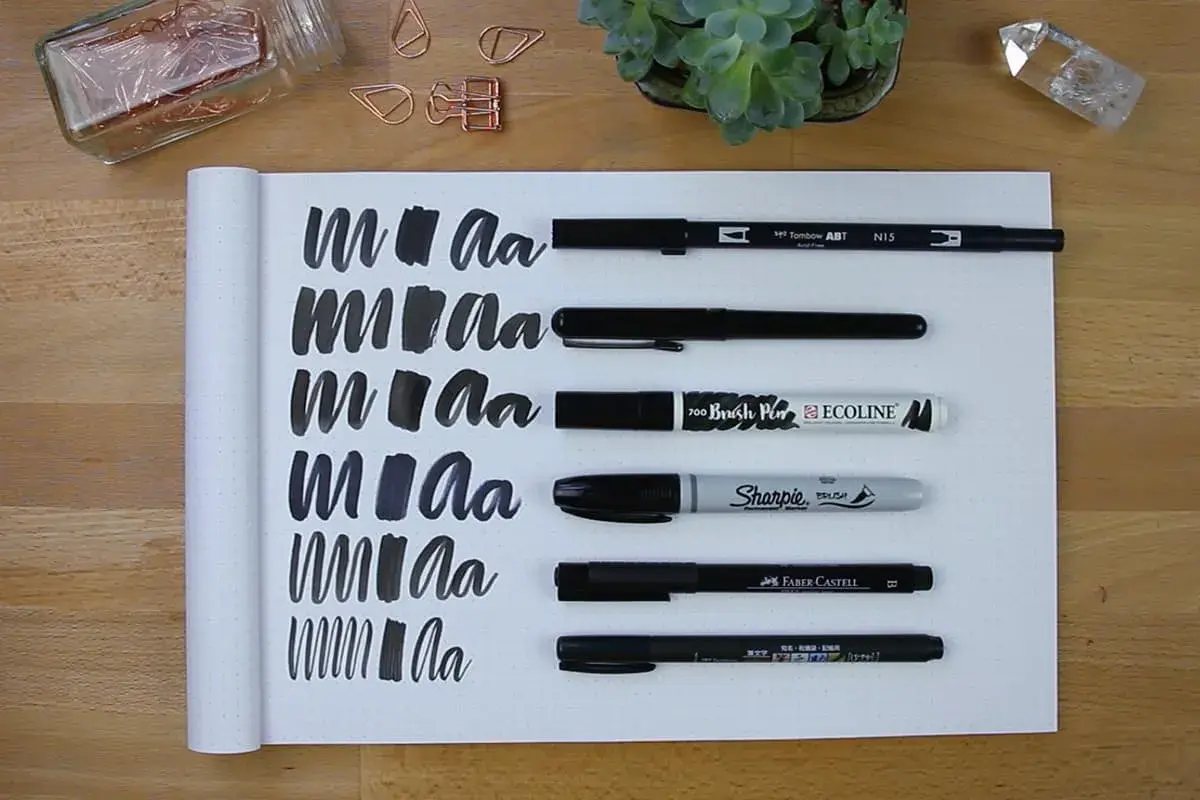 Práctica de lettering con varios rotuladores, mostrando trazos y letras "m" y "a" en un cuaderno.