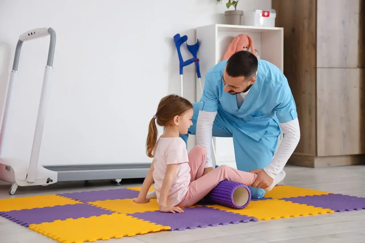 Fizjoterapeuta stosuje metodę Bobath podczas ćwiczeń z dziewczynką, która leży na macie z wałkiem pod nogą.