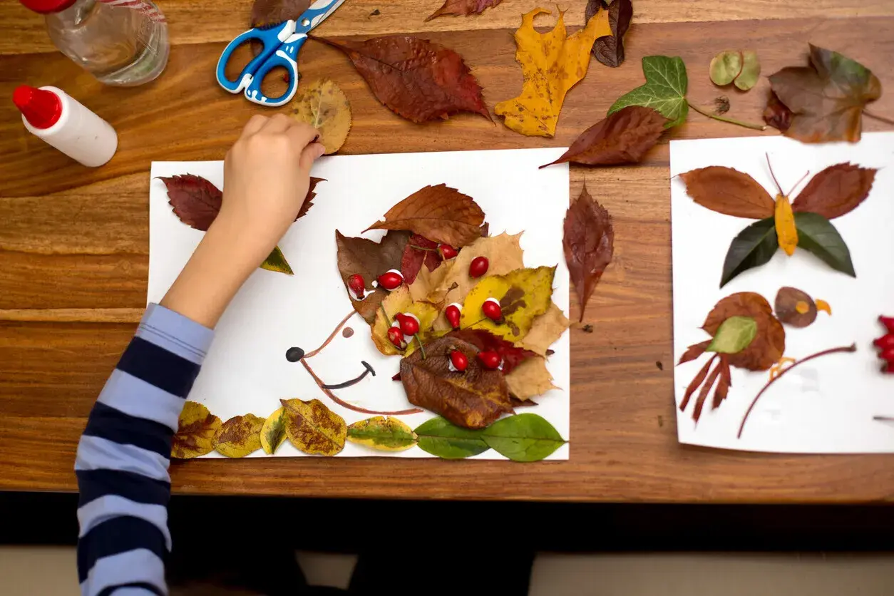 Niño haciendo otoño manualidades preescolar: un erizo con hojas secas y bayas rojas, junto a una mariposa y un pájaro hechos de hojas.