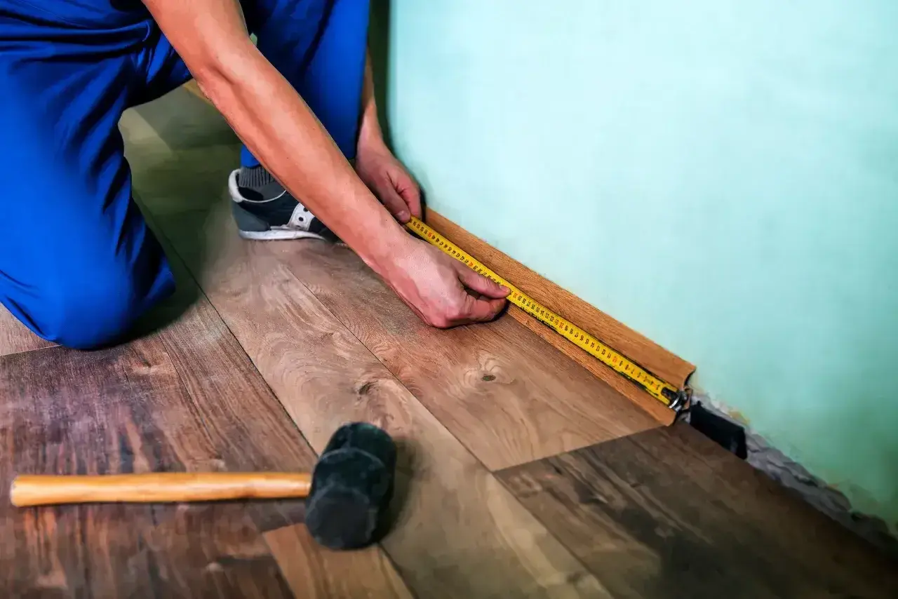 Mężczyzna w niebieskich spodniach mierzy taśmą podłogę, przygotowując się do tego, jak kłaść panele. Obok leży gumowy młotek.