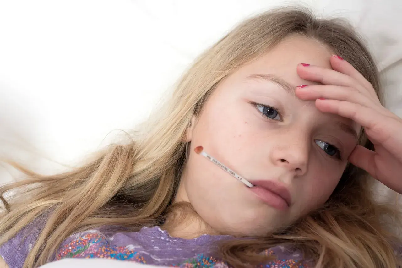 Une jeune fille malade avec un thermomètre dans la bouche, se demandant combien de temps dure la mononucléose.