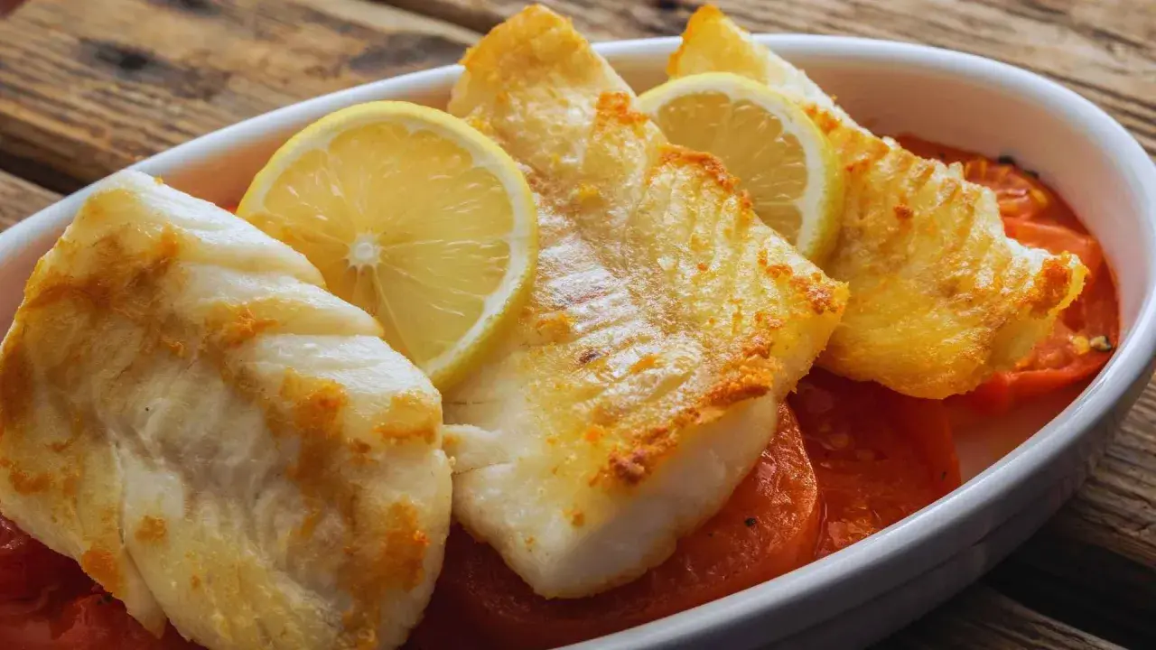 Filetes de pescado blanco horneado con rodajas de limón y tomates en una fuente blanca.