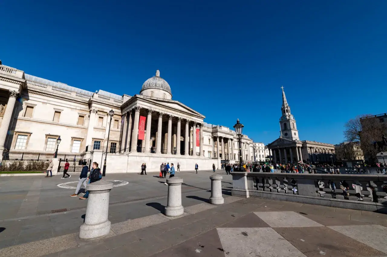 Trafalgar Square w Londynie, z National Gallery w tle. Wiele osób spaceruje po placu, ciesząc się darmowymi muzeami Londyn.