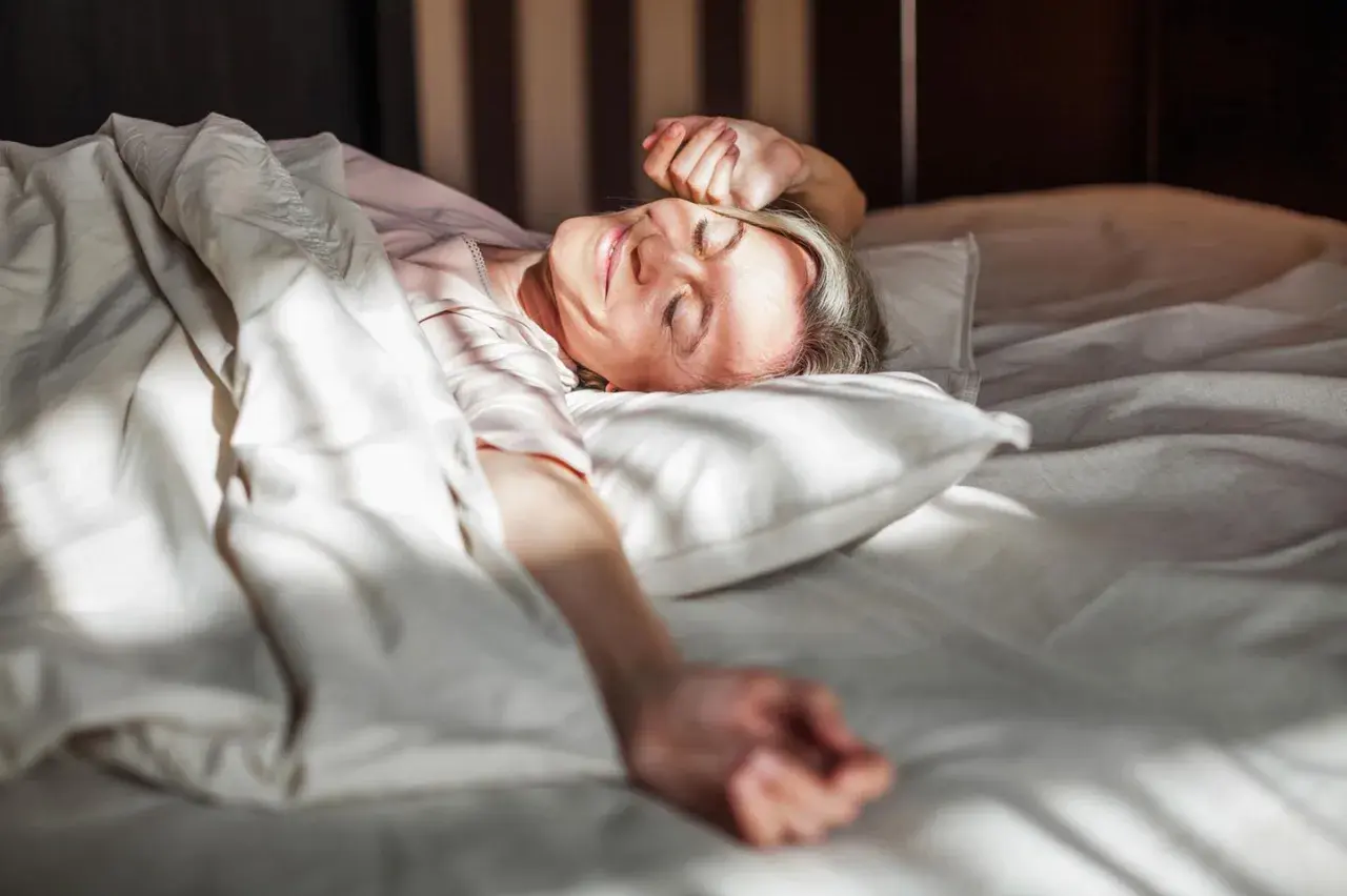 Frau erwacht lächelnd im Bett, genießt die Sonne. Erfüllt die mindest schlafzeit für Wohlbefinden.
