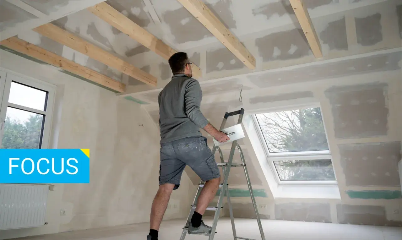 Un operaio su una scala lavora al soffitto, applicando stucco su pannelli di fibrogesso, materiale versatile per ristrutturazioni.