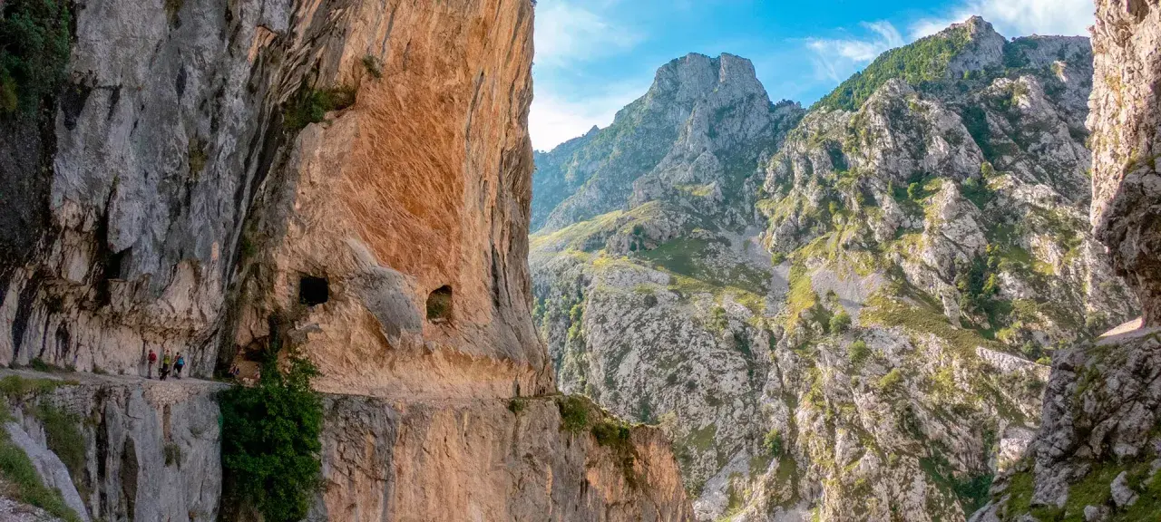 Senderos tallados en roca, con personas caminando. El inicio y fin de la ruta del Cares se pierden entre imponentes montañas verdes y cielos azules.