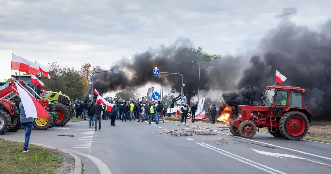 Czerwony traktor na drodze, w tle dym i ludzie z polskimi flagami.