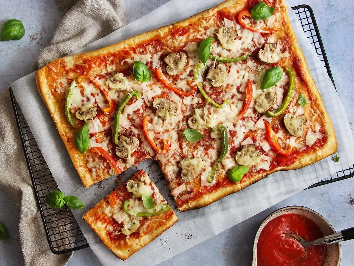 En avskuren bit av en rektangulär pizza med skinka, champinjoner och paprika. Vilken skinka till pizza är bäst? Frågan är svår att svara på, men denna ser god ut!