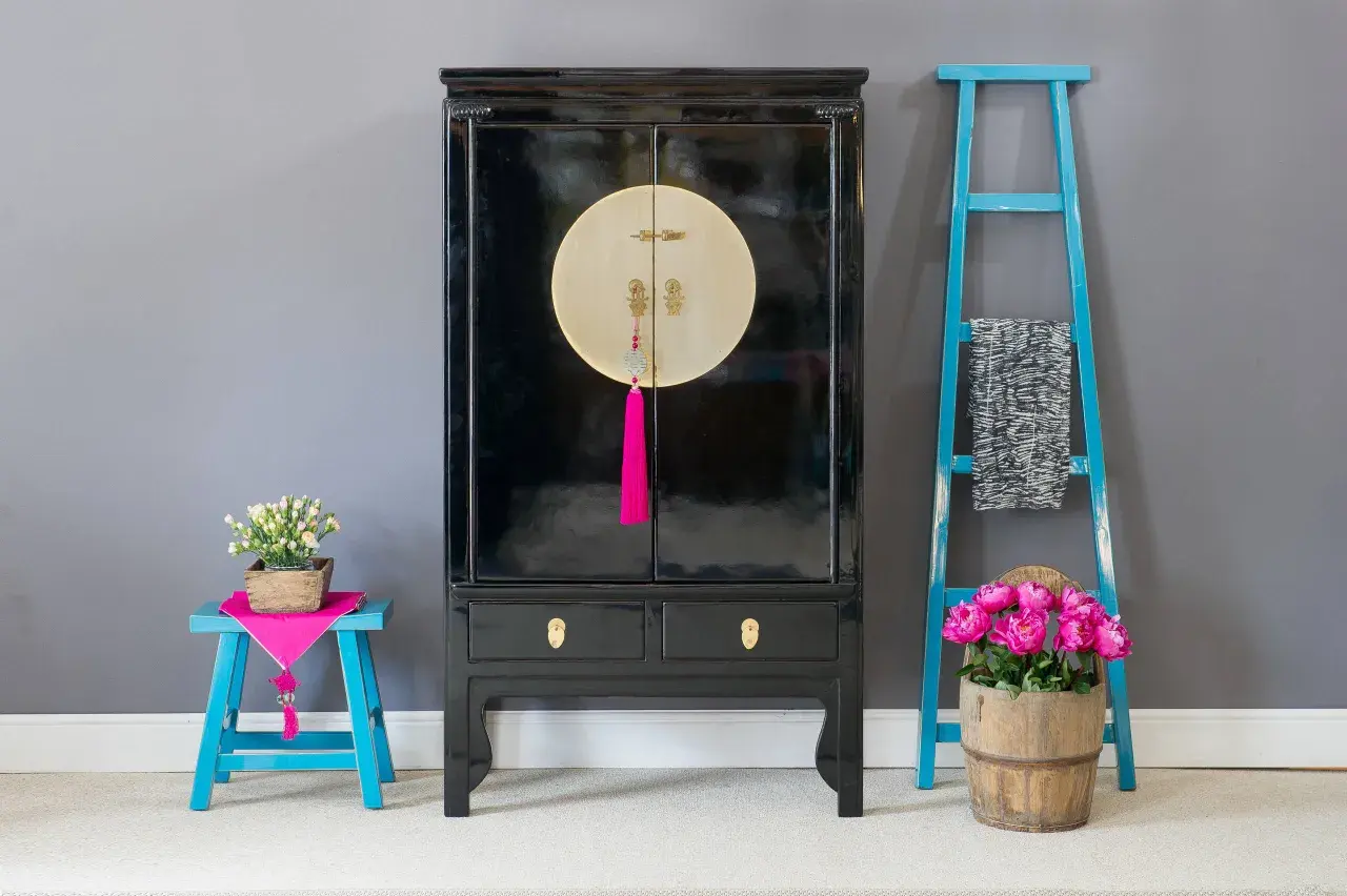 Mobile con laccatura nera lucida, dettagli dorati e nappa fucsia. Accanto, sgabello blu con fiori e scaletta decorativa.