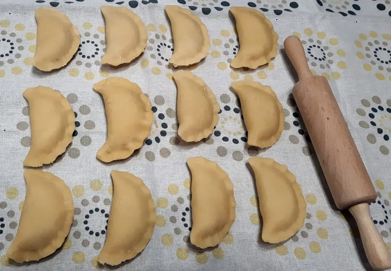 Gotowe do gotowania pierogi z jabłkami, ułożone rzędami na wzorzystym obrusie, obok drewnianego wałka.