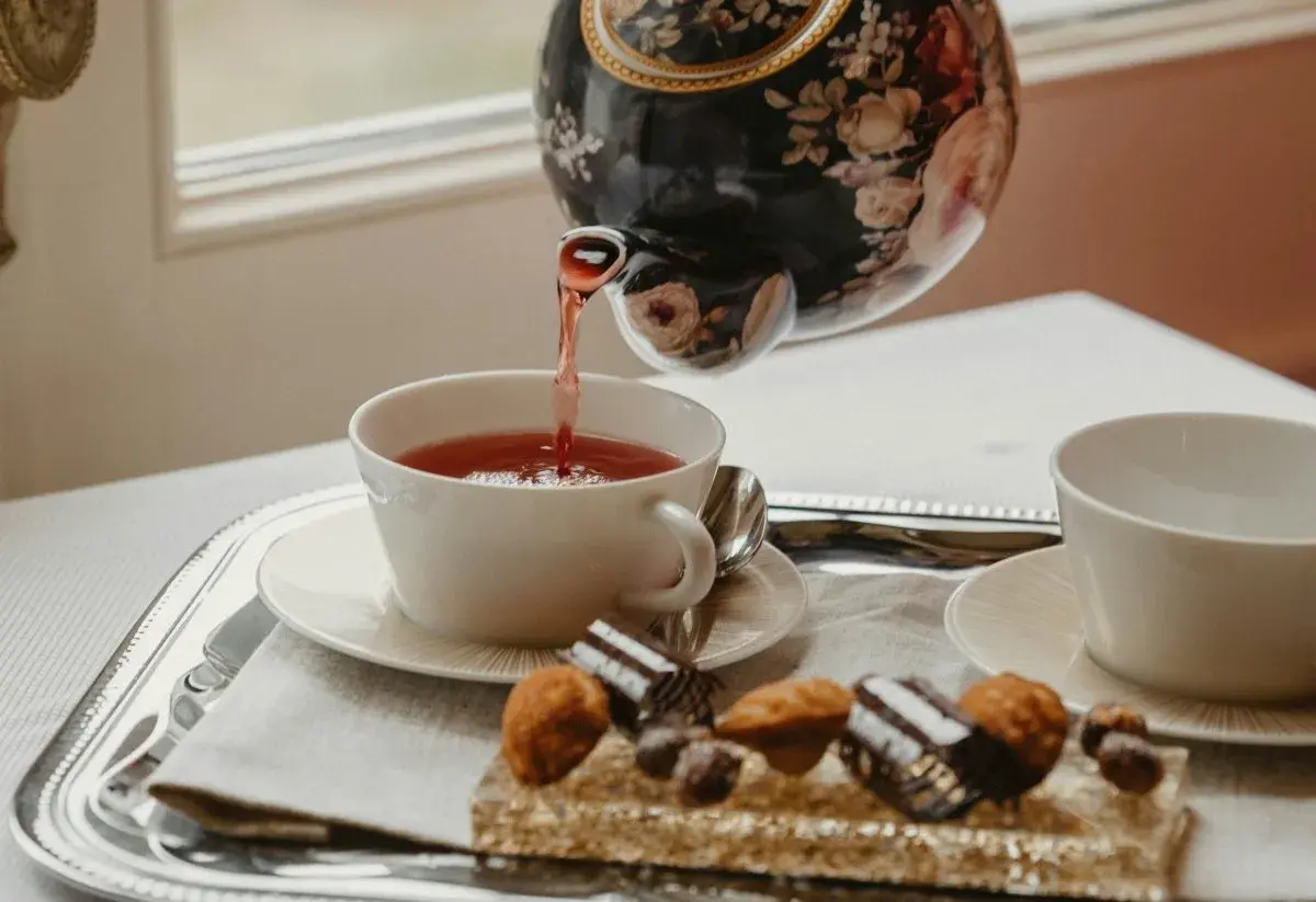Tetera floral vierte té rojo en taza blanca. Bandeja con dulces y otra taza, perfecto para la hora del té en Inglaterra.