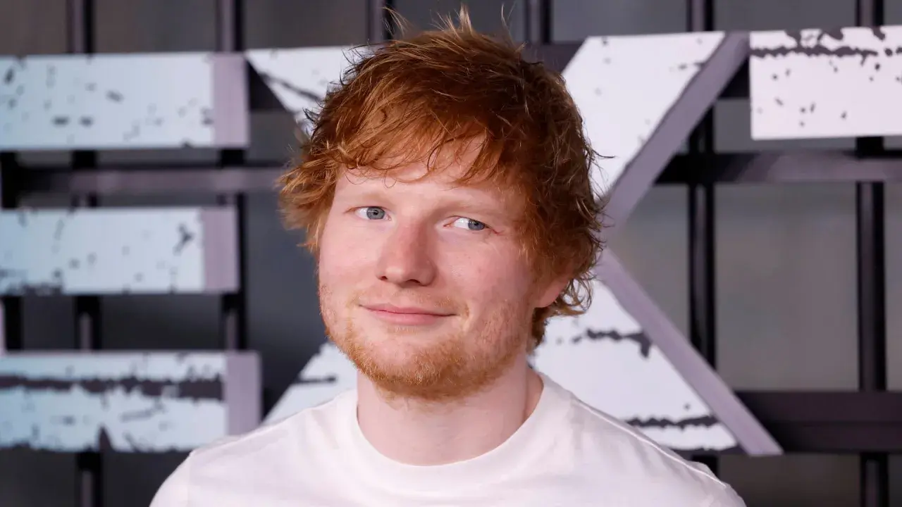 Ed Sheeran w białej koszulce, z rudymi włosami i niebieskimi oczami, uśmiecha się.