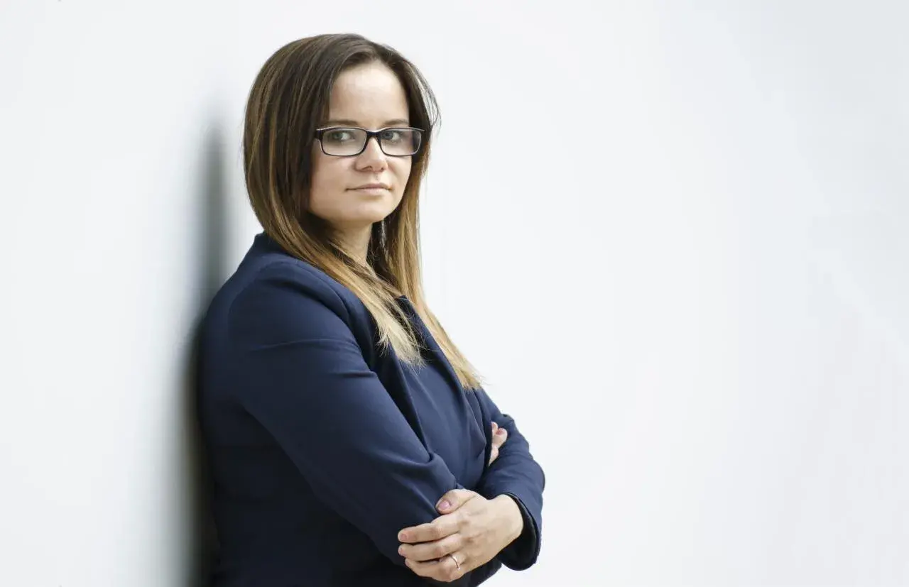Kobieta w okularach i granatowej marynarce, oparta o białą ścianę.