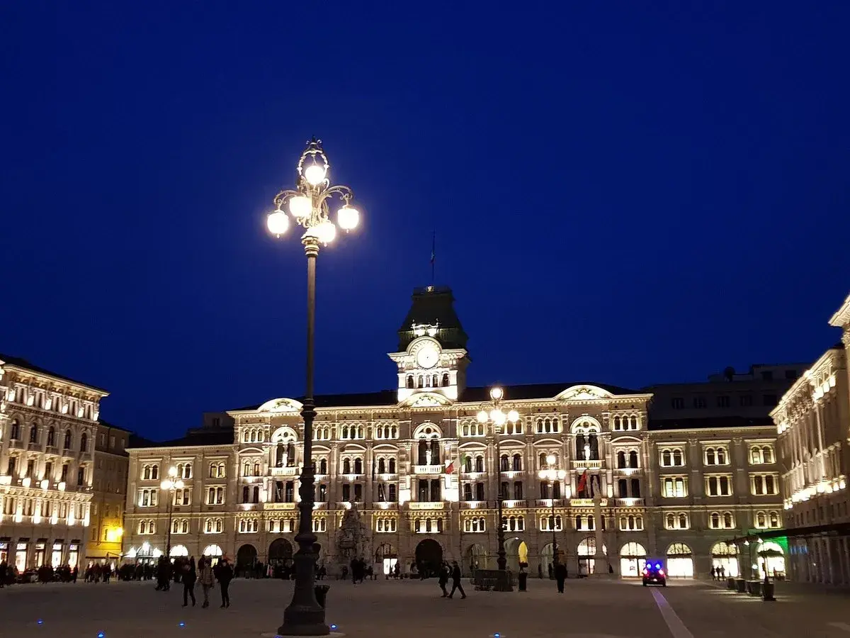 Piazza Unità d'Italia a Trieste, illuminata di sera. Edifici storici e lampioni creano un'atmosfera magica.
