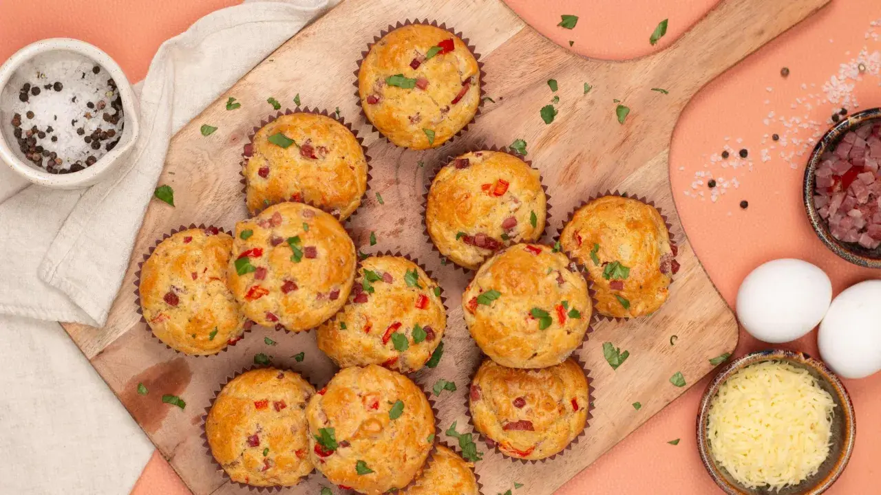 Herzhafte Pizza Muffins mit Speck und Paprika, garniert mit Petersilie. Daneben Zutaten wie Eier, Käse und Gewürze.