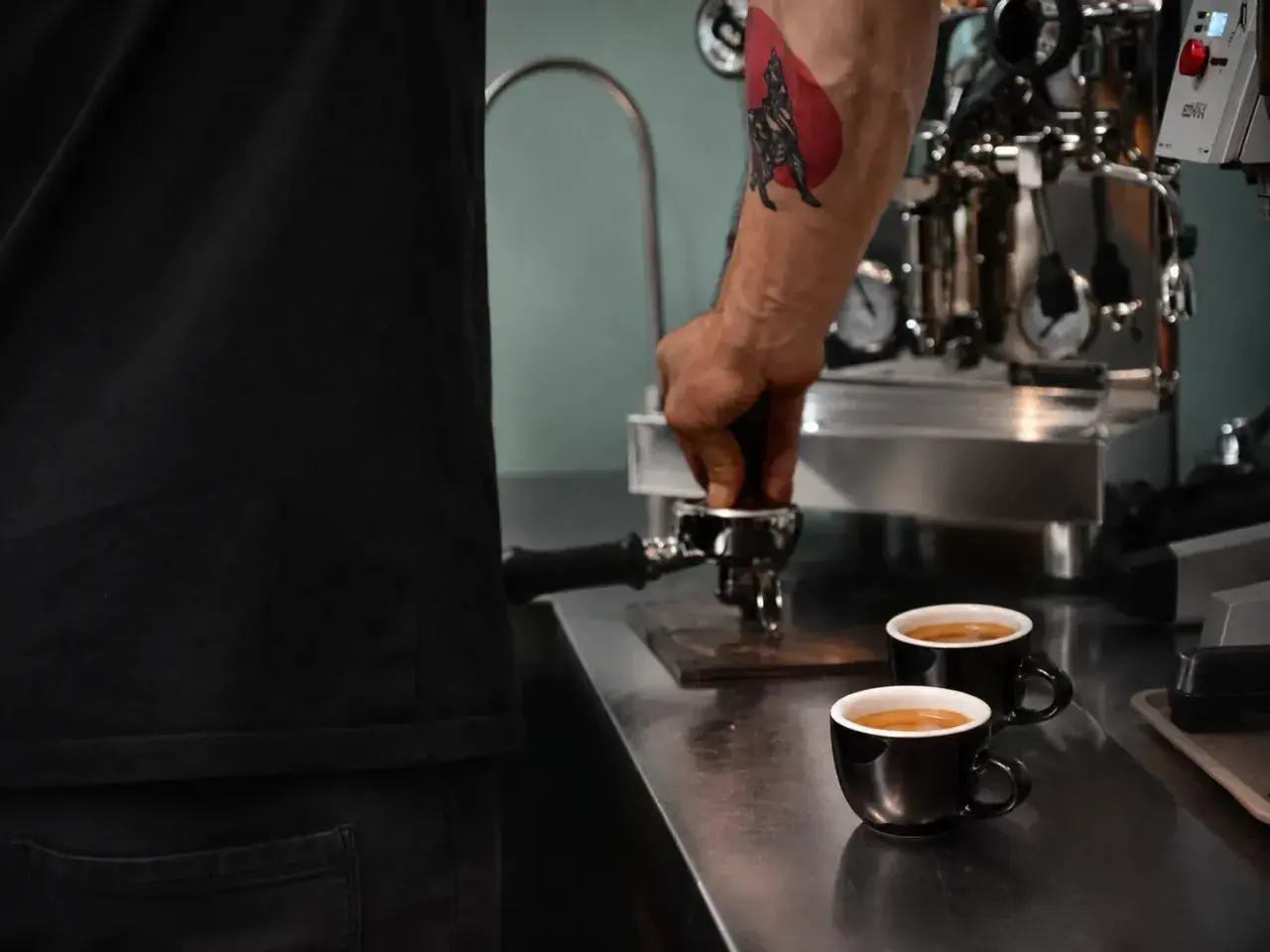 Barista z tatuażem na ramieniu przygotowuje kawę americana. Dwie filiżanki czarnej kawy stoją na blacie obok ekspresu.