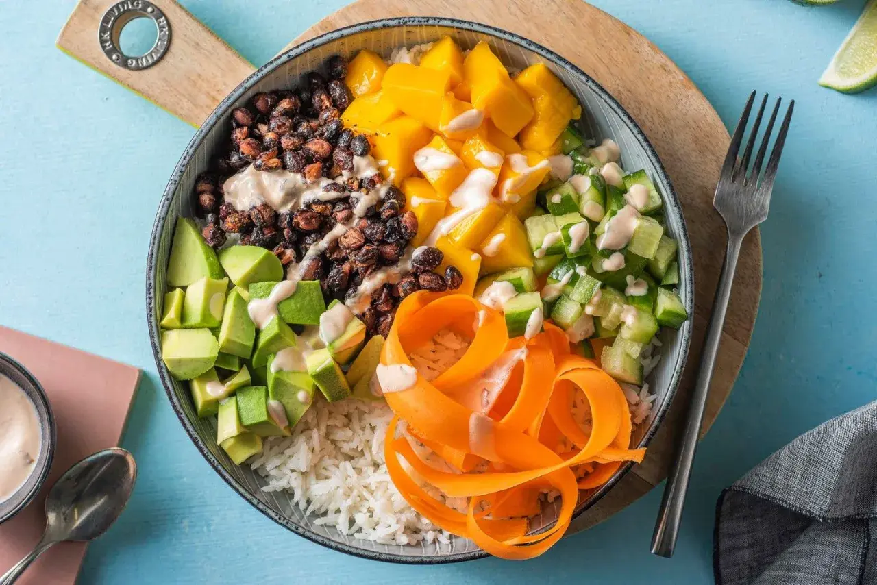 En färgglad poke bowl med ris, svarta bönor, mango, gurka, avokado och morotsstrimlor, toppad med en krämig dressing.