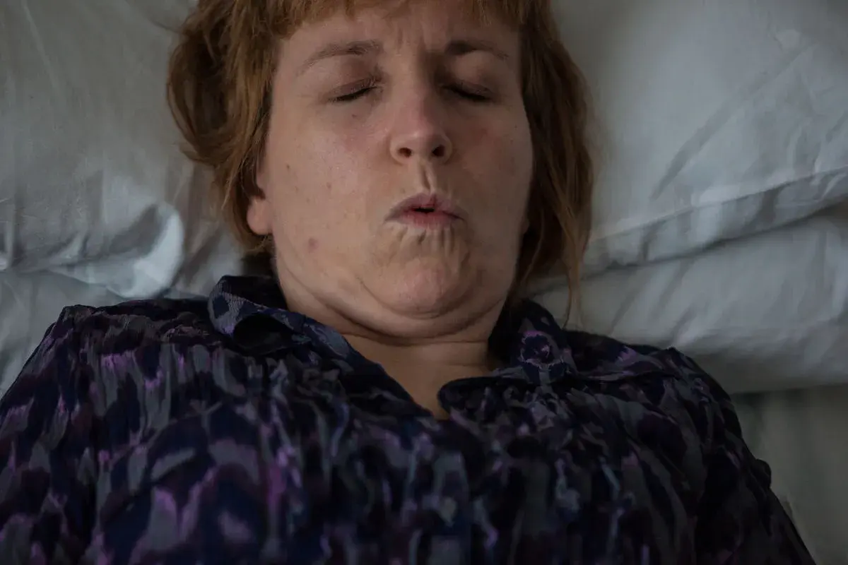 Donna con occhi chiusi e bocca socchiusa, forse a causa di una disestesia neurologica. Indossa una camicetta viola.