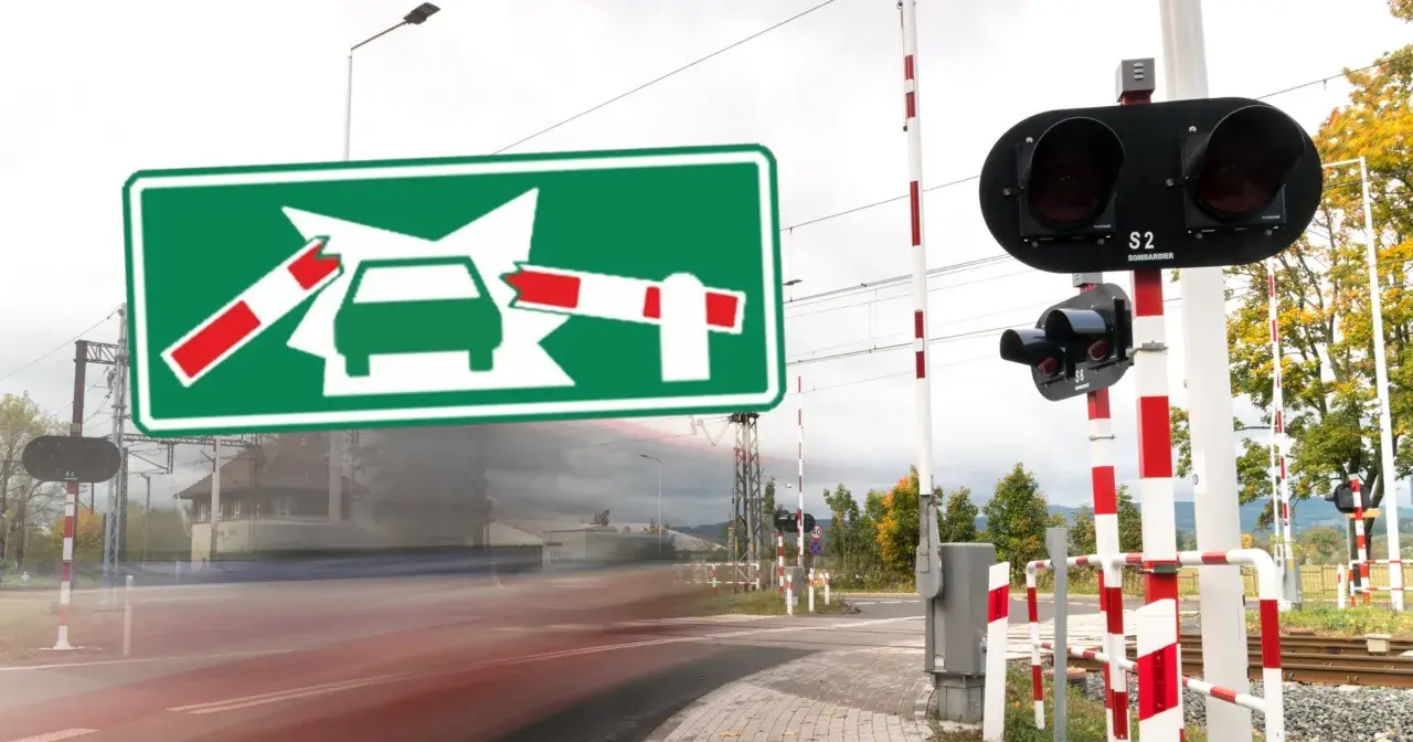 Zielony znak informuje o oznaczeniu przejazdu kolejowego. Po bokach widać rogatki i sygnalizację świetlną.