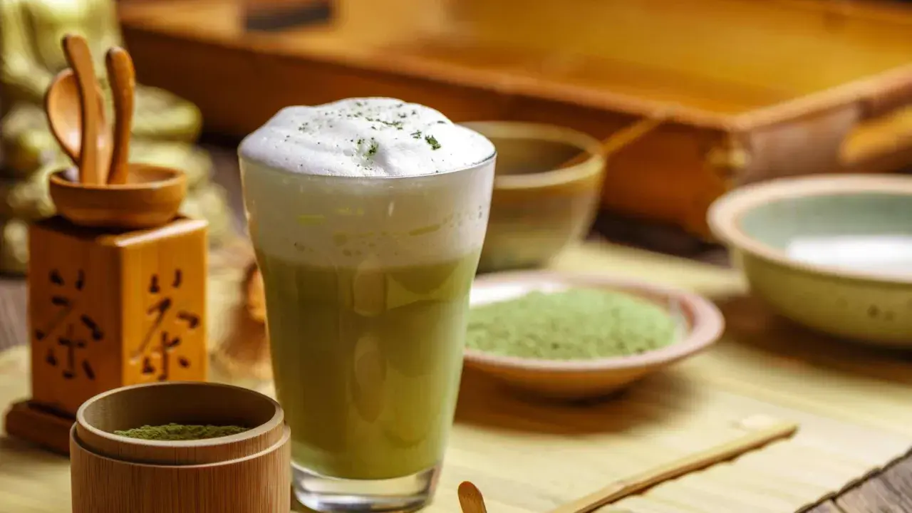 Ein cremiger Matcha Latte mit Milchschaum, umgeben von Utensilien für die Matcha Tee Zubereitung.