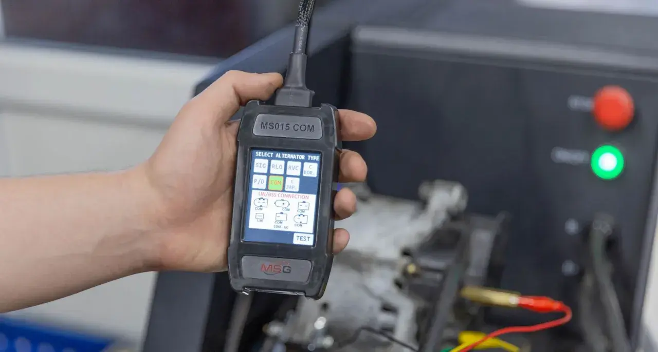 Dłoń trzyma tester diagnostyczny do alternator w samochodzie. Ekran pokazuje wybór typu alternatora i połączeń LIN/BSS.