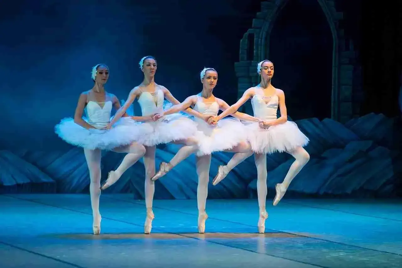 Quattro ballerine in tutù bianchi eseguono una danza aggraziata sul palco, con un arco di pietra sullo sfondo.