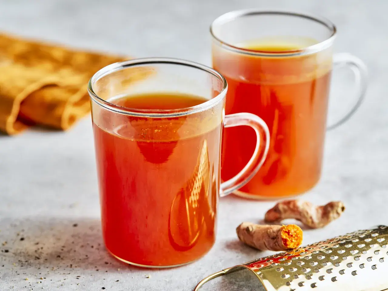 Zwei Gläser mit leuchtend orangefarbenem Getränk, das nach einem Kurkuma Honig Rezept zubereitet wurde. Frische Kurkumawurzeln und ein Sieb liegen daneben.