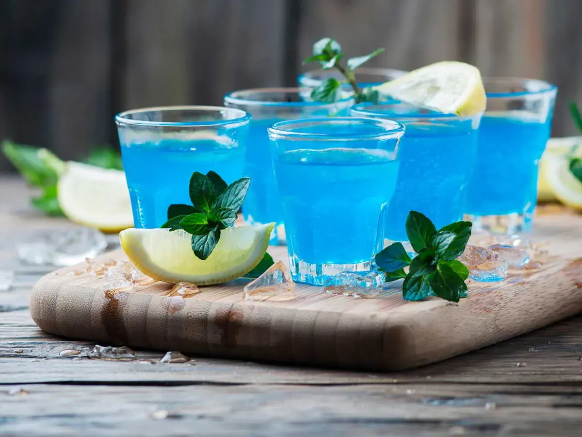 Cocktail martini blanc curaçao, bleu vif, garni de menthe fraîche et de quartiers de citron, sur un plateau en bois avec de la glace pilée.