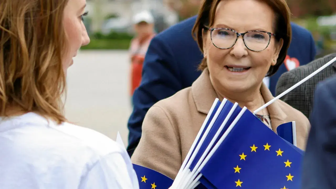Ewa Kopacz, gdzie mieszka, uśmiecha się, trzymając flagi UE.