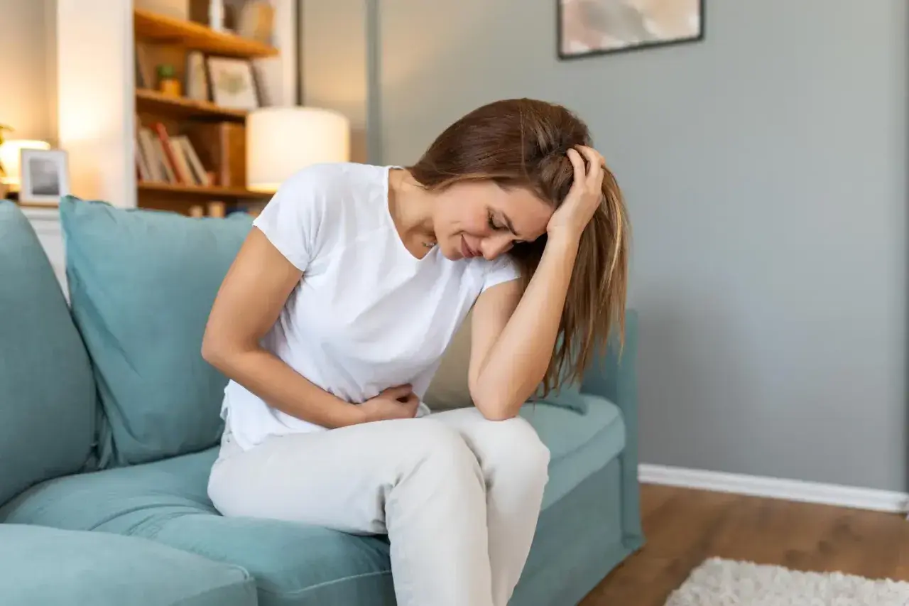 Femme assise sur un canapé bleu, se tenant le ventre et la tête, visiblement souffrante. Elle pourrait exprimer la douleur liée à l'endométriose profonde.