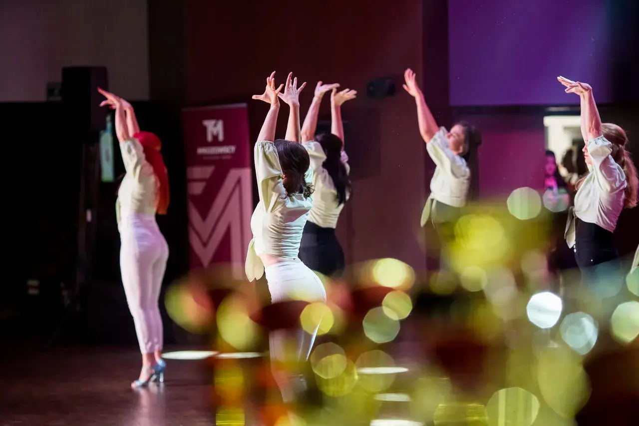 Tancerki w jasnych bluzkach i białych spodniach wykonują zmysłowy taniec. W tle logo szkoły tańca. To piękna **bachata solo**.